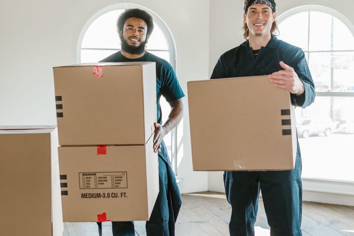 Two movers carrying cardboard boxes through a bright room