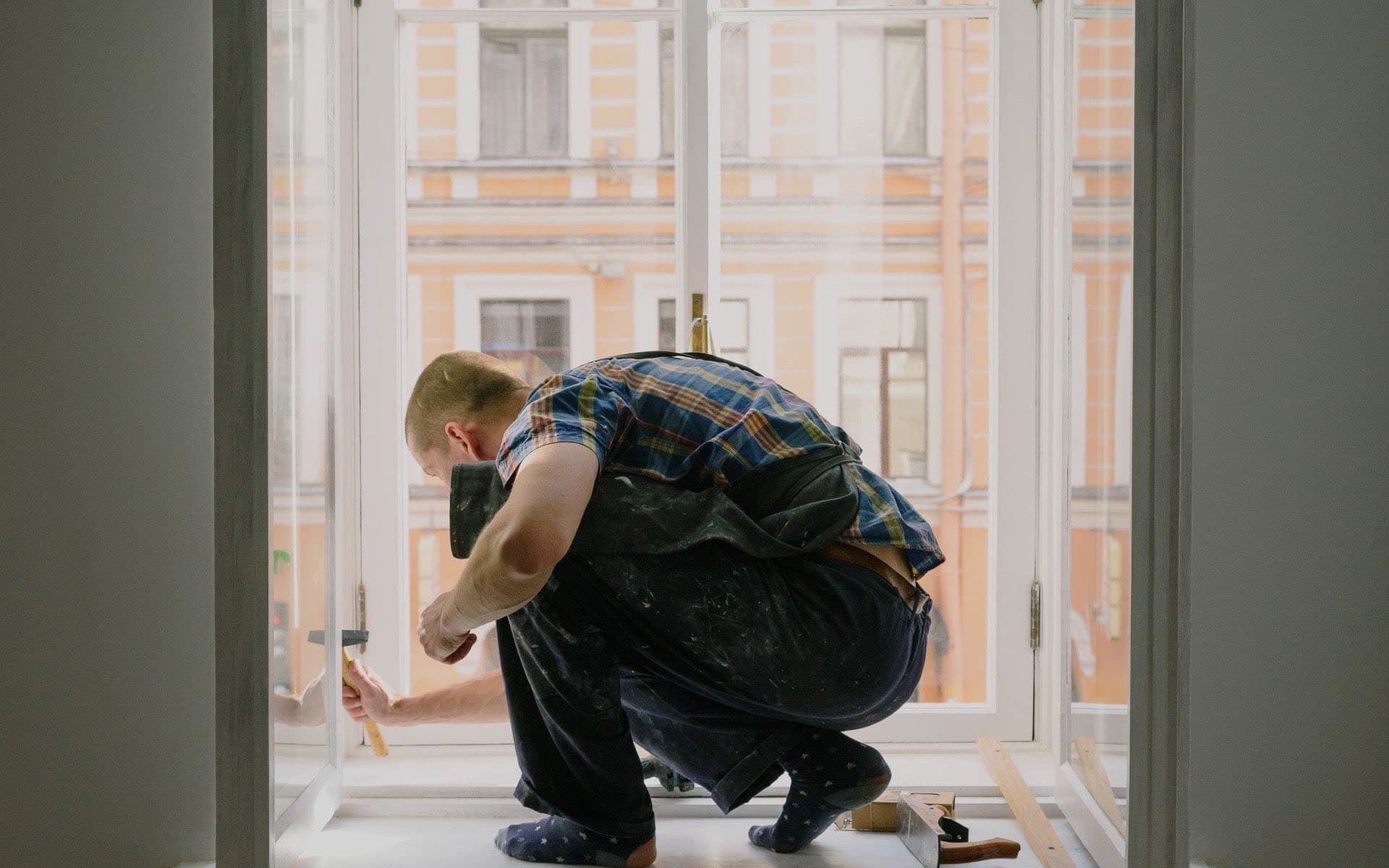 A technician crouched on a windowsill, repairing the window frame with a hammer